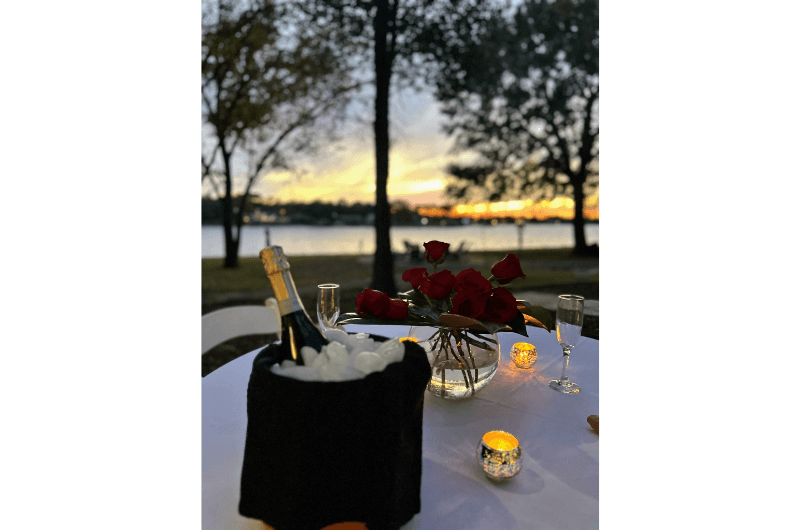 A romantic table setting by a lake at sunset, featuring champagne, roses, and candles.