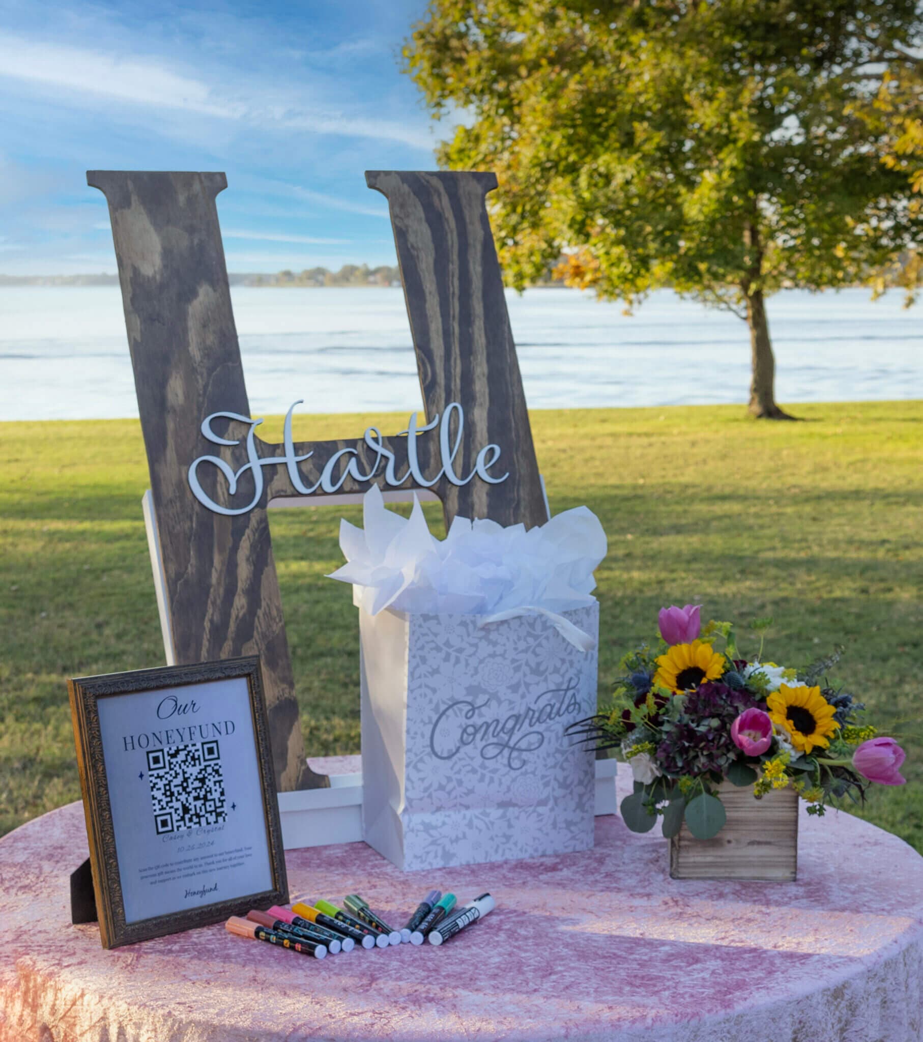A decorative wedding display featuring a "Hartle" sign, a gift box, a honeyfund sign with a QR code, and a floral arrangement by a waterfront.