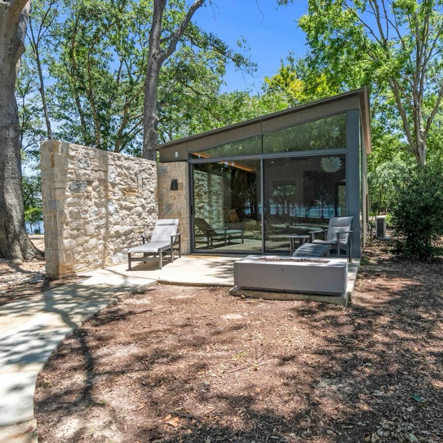 Modern cabin with glass walls nestled among trees, featuring patio seating and a stone wall.
