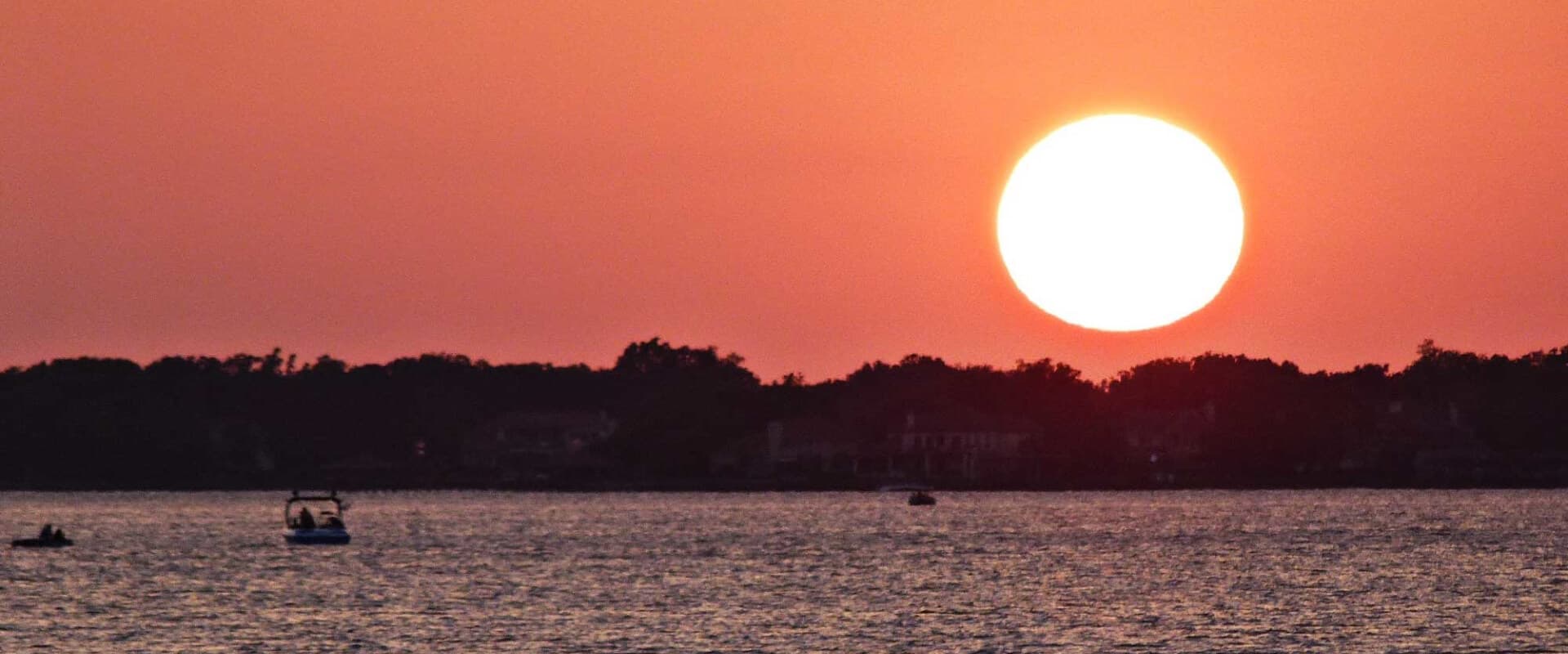 A large orange sun sets over a calm waterway, with silhouettes of boats and trees in the foreground.