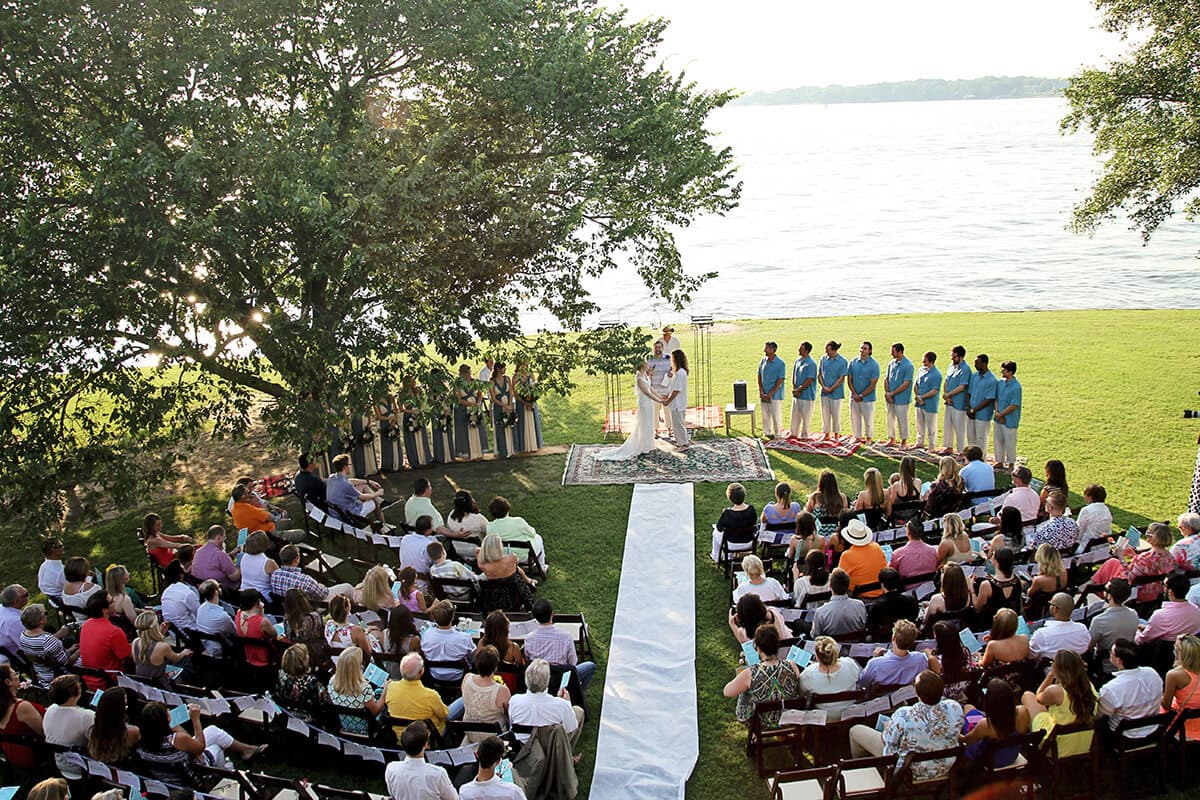 A wedding ceremony takes place outdoors by a lake, with guests seated on either side of a white aisle.
