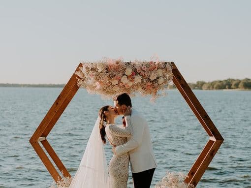 A newlywed couple shares a kiss under a floral arch by the water.