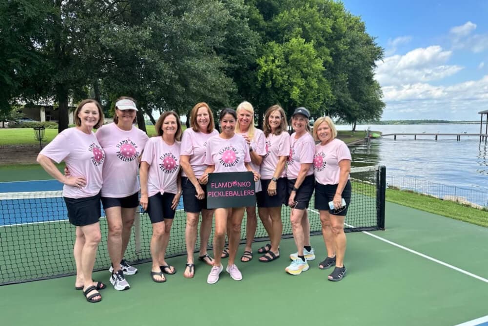 A group of nine women wearing matching pink t-shirts poses together on a pickleball court by a lake.
