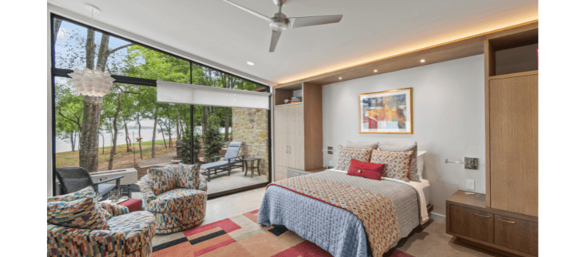 A modern bedroom with a large window showcasing a view of greenery, featuring a neatly made bed and colorful armchairs.