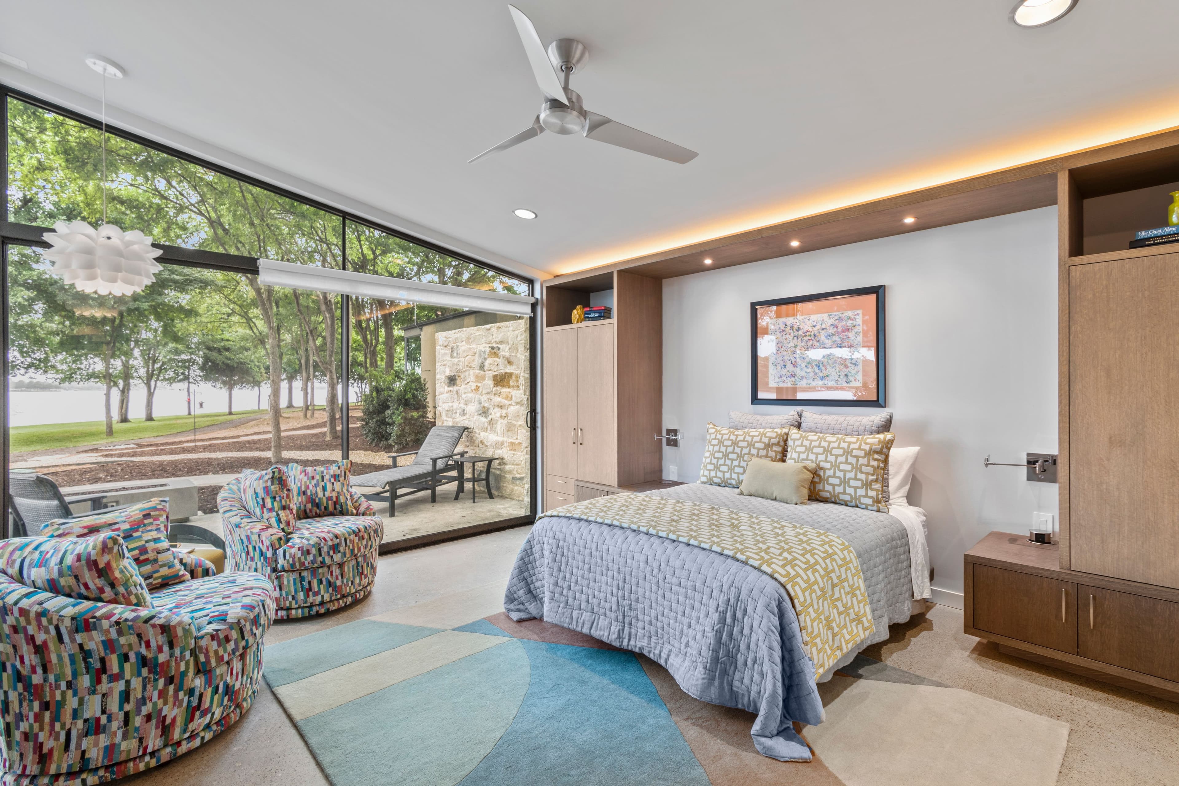 A modern bedroom with large windows, featuring a bed, colorful chairs, and natural light.