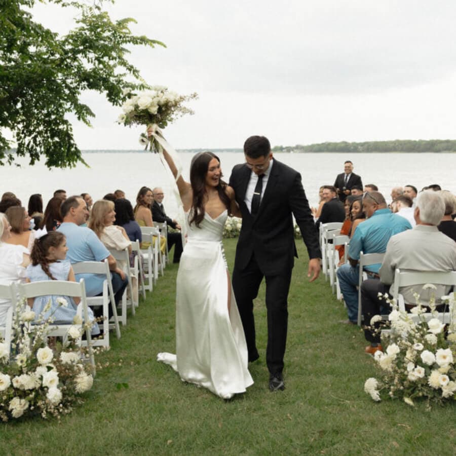 A newlywed couple joyfully walks down the aisle, surrounded by guests, by a lakeside setting.