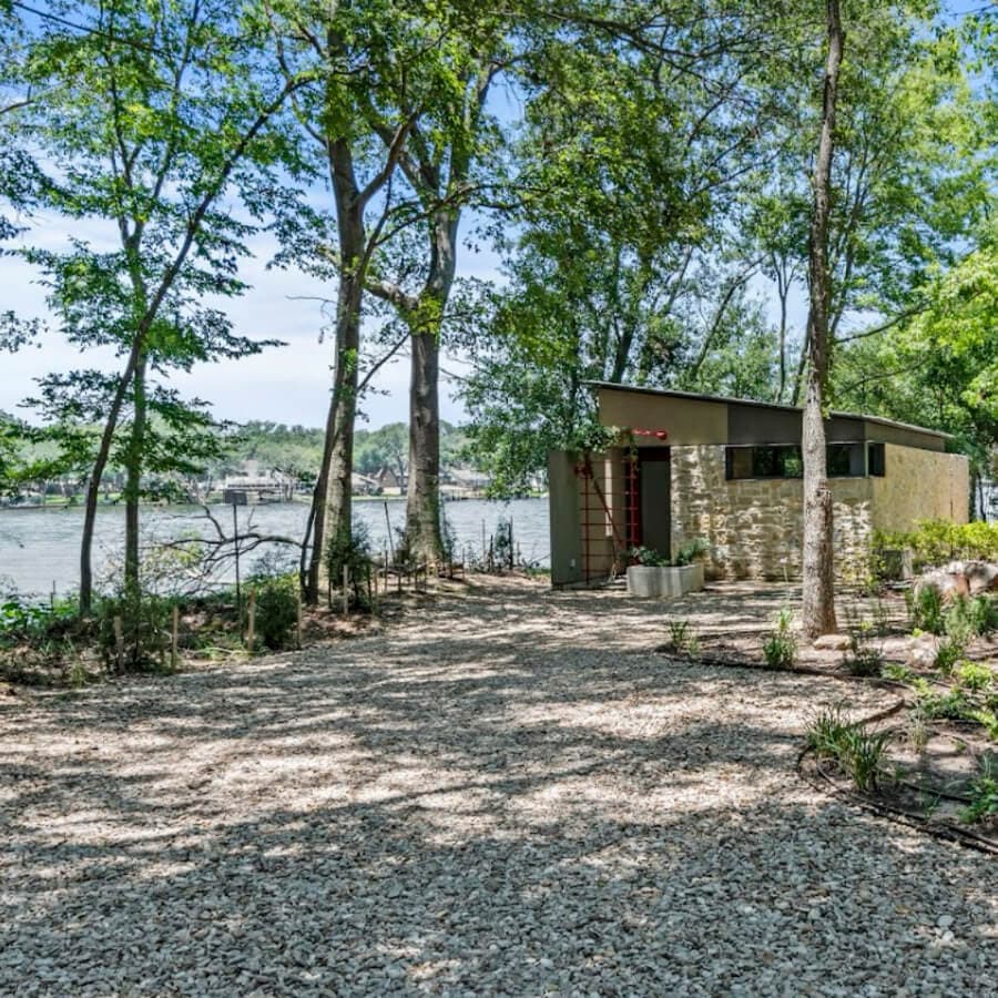 A modern cabin sits among trees near a river, surrounded by gravel pathways.