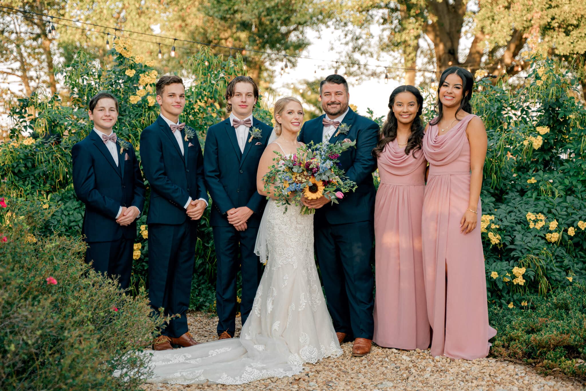 A wedding party poses together outdoors, featuring the bride in a white gown and the groom in a dark suit, surrounded by bouquets and floral decorations.
