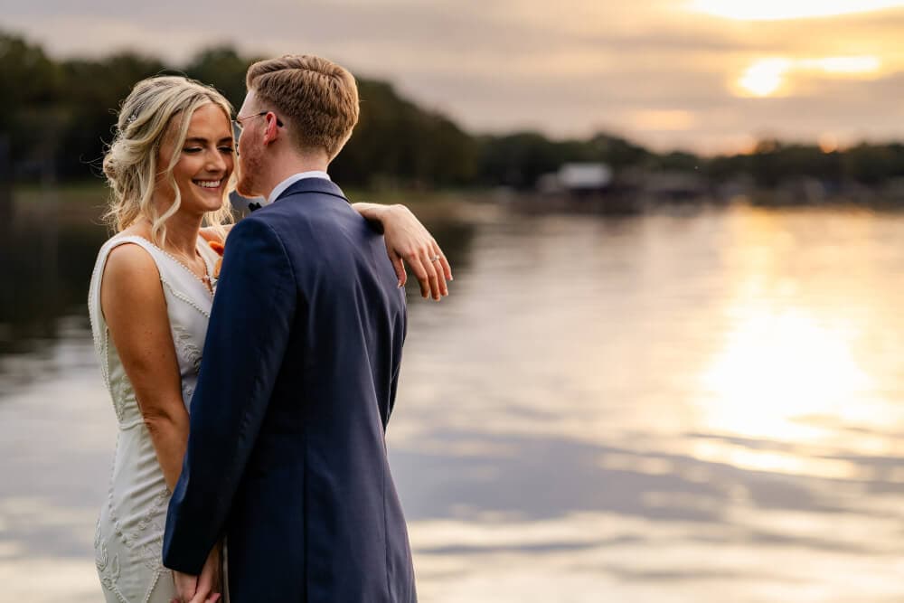 A smiling couple embraces by a serene lake at sunset.