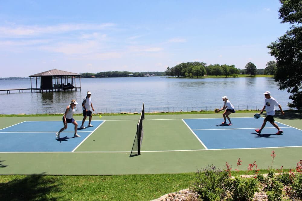 Four people play pickleball on a lakeside court under a clear blue sky.