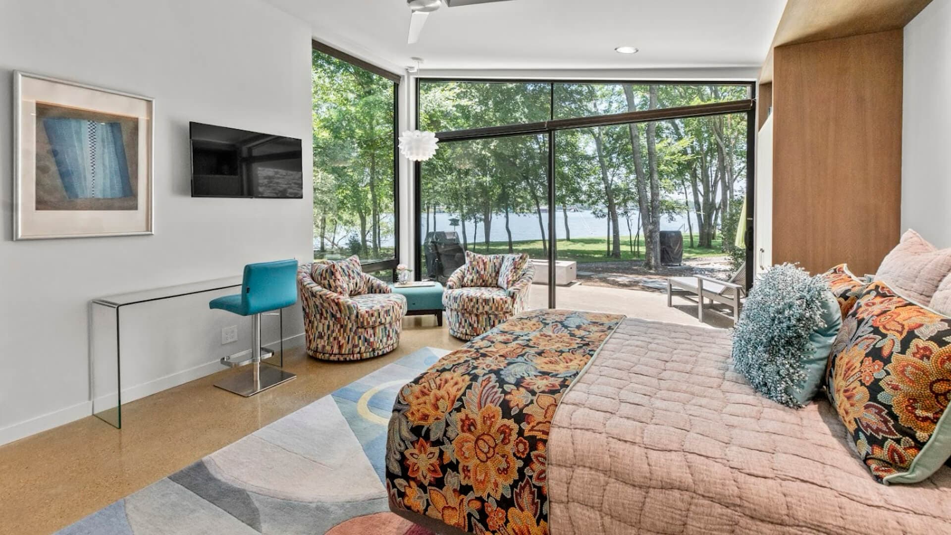 A modern bedroom with large windows overlooking a lake, featuring a colorful bedspread, two armchairs, and a small desk.