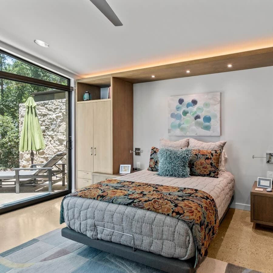 A modern bedroom featuring a bed with floral bedding, a chair, and large glass doors leading to an outdoor area with a green umbrella.
