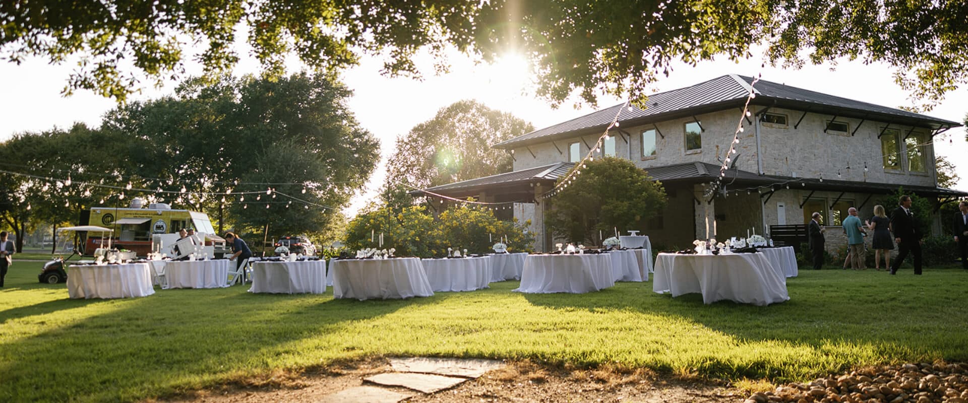Outdoor event setup with tables under string lights and a sunlit house in the background.