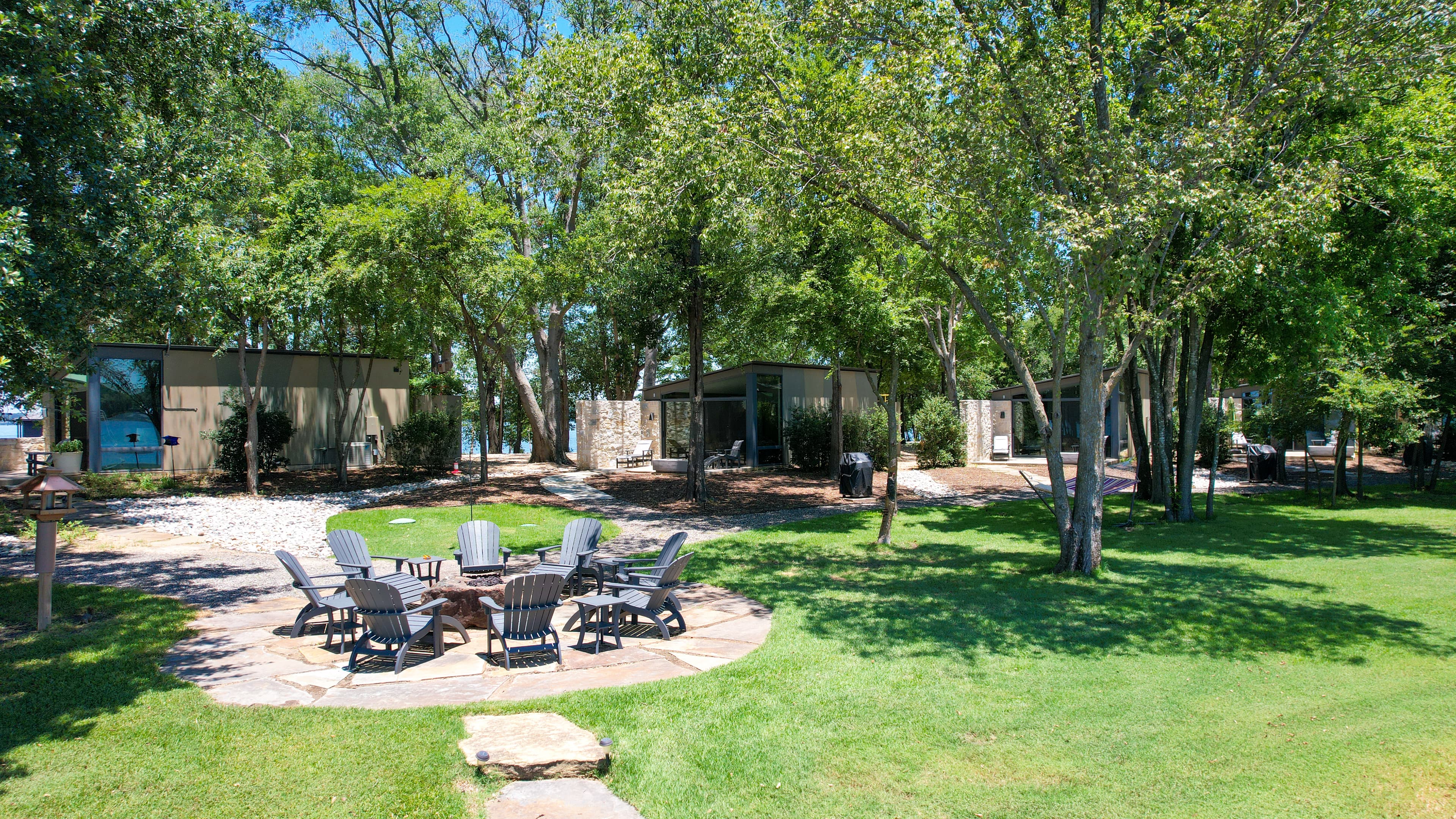 Stone patio with a central fire pit surrounded by lounge chairs on a grassy lawn shaded by trees