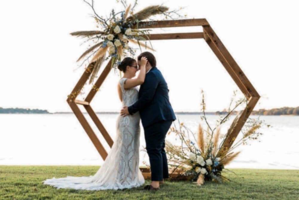 A couple kisses under a floral arch by the water at their wedding.