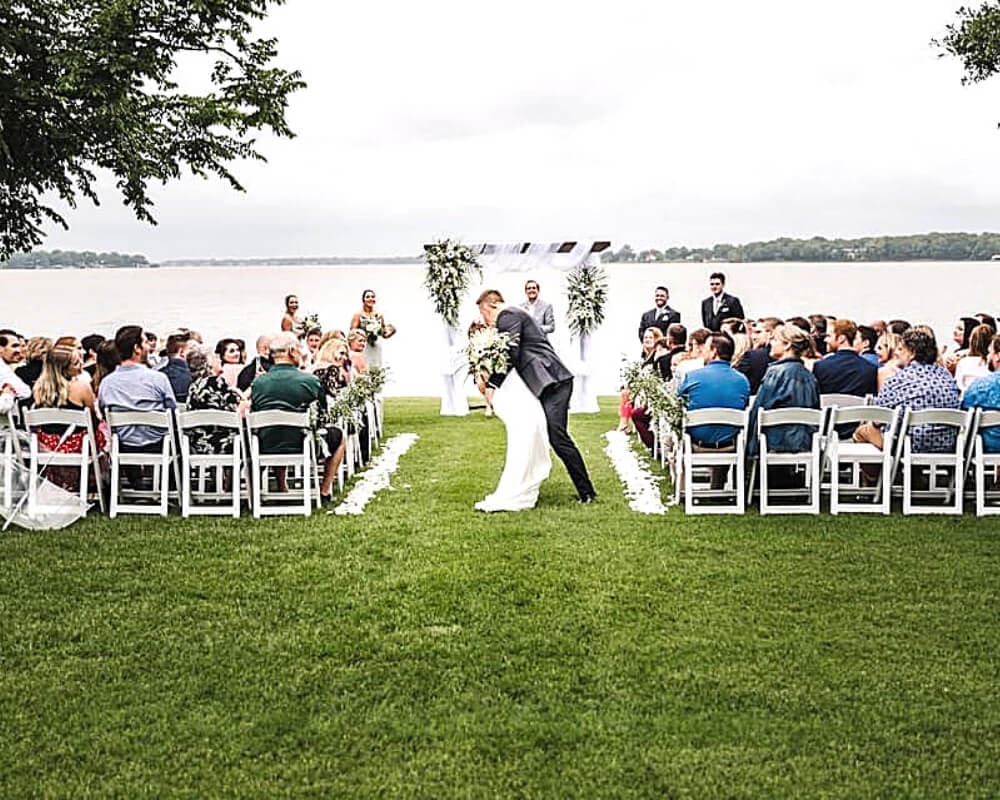 A couple shares a kiss during their wedding ceremony by a lakeside, surrounded by guests.
