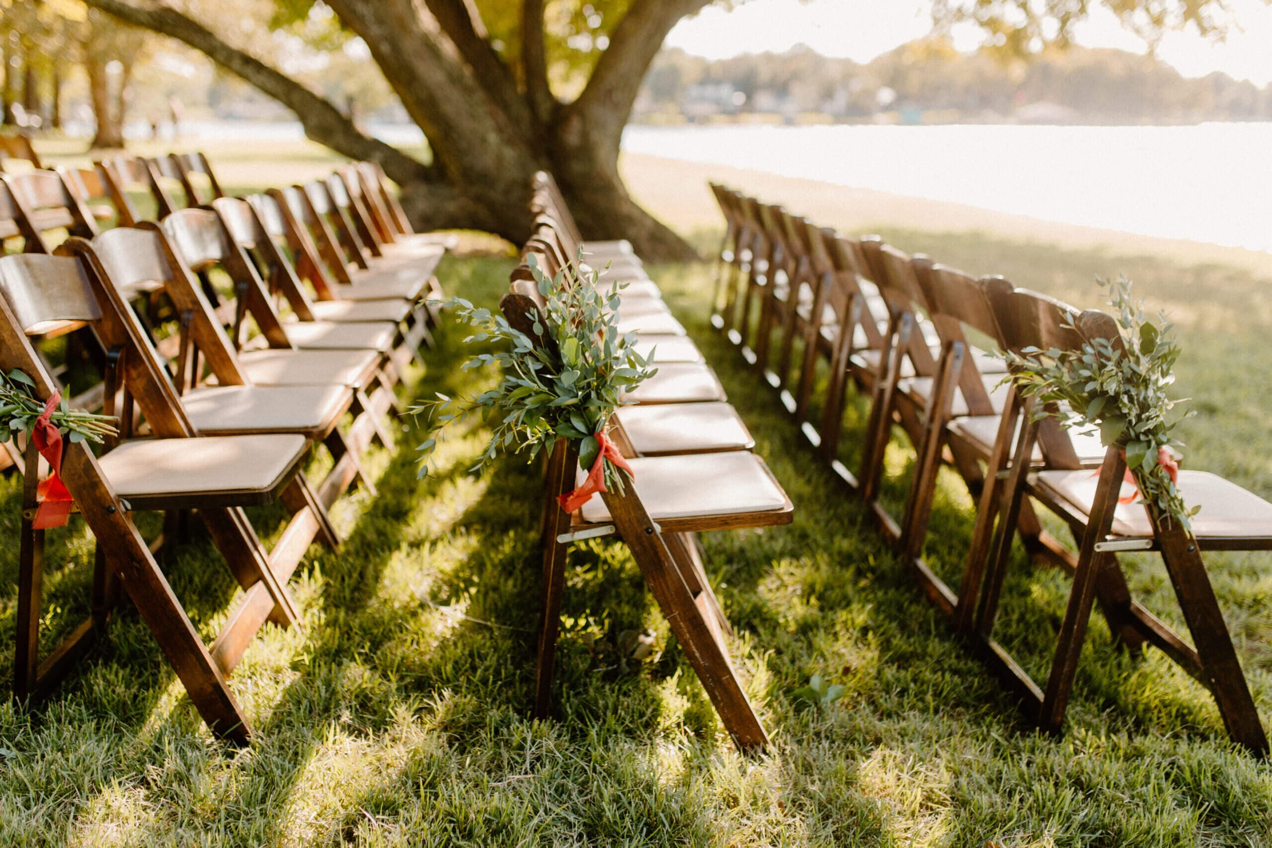 Wooden chairs arranged for an outdoor event, adorned with greenery and ribbons under a tree by a waterfront.