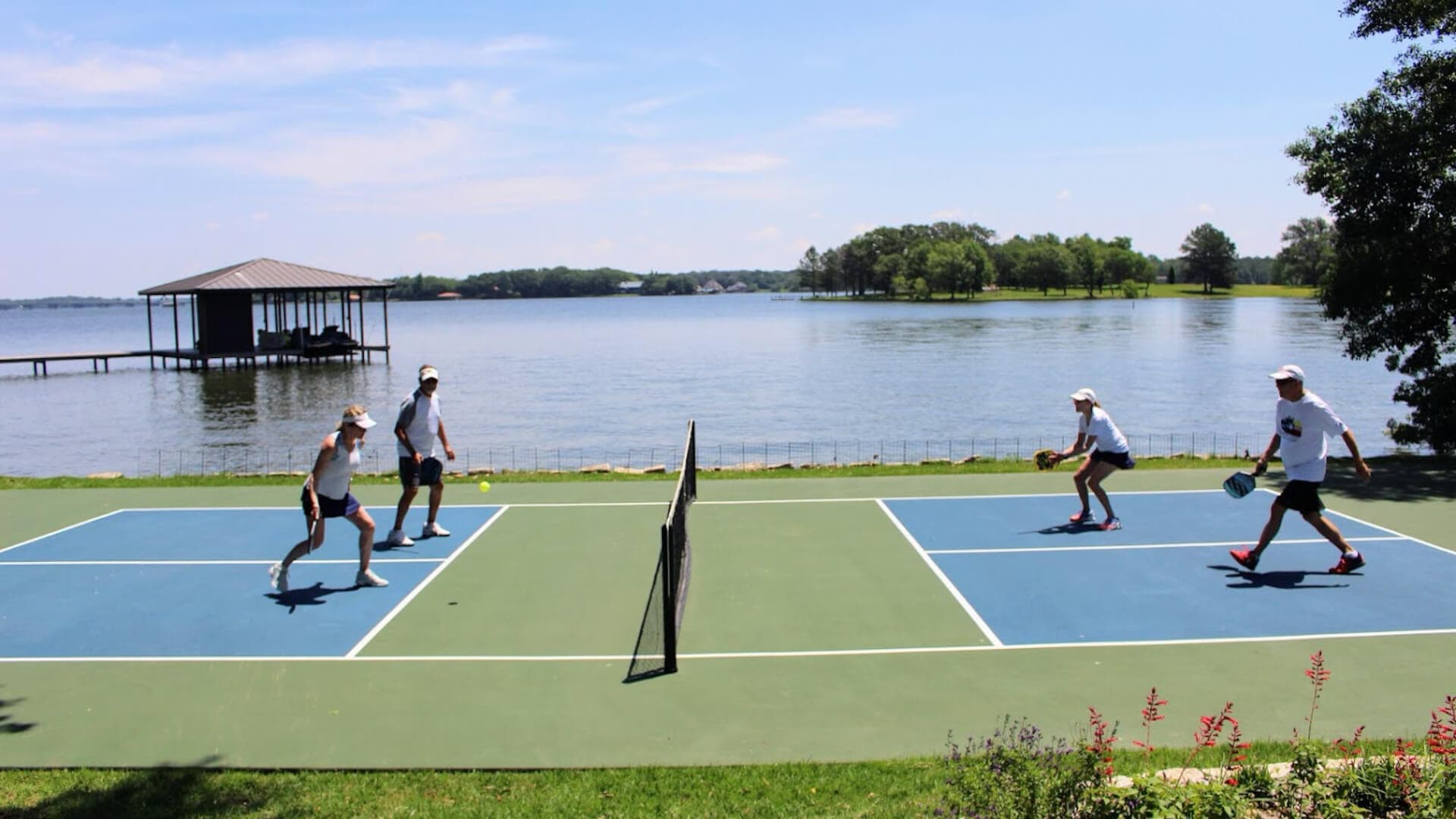 Four people play pickleball on a court by a lake under a clear sky.