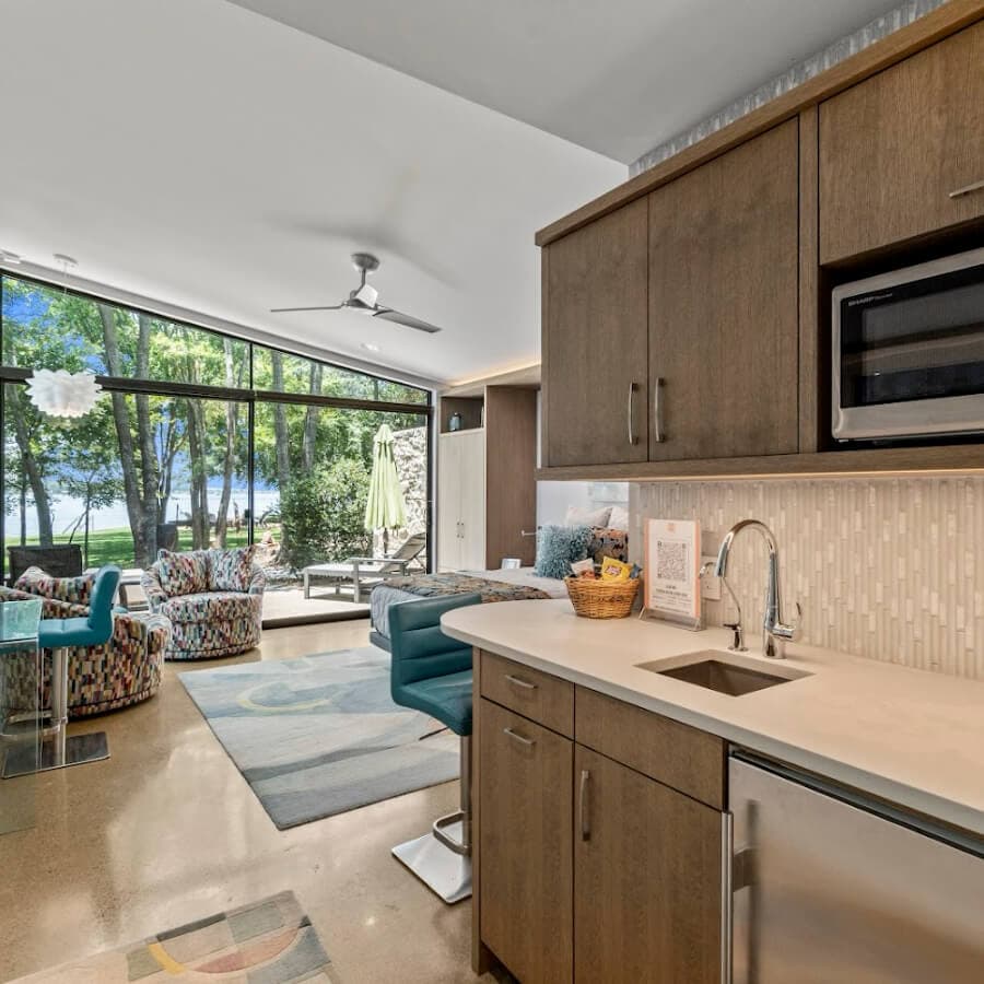 A modern kitchen area featuring wooden cabinets and a sink, opening up to a bright living space with colorful seating and large windows overlooking greenery.