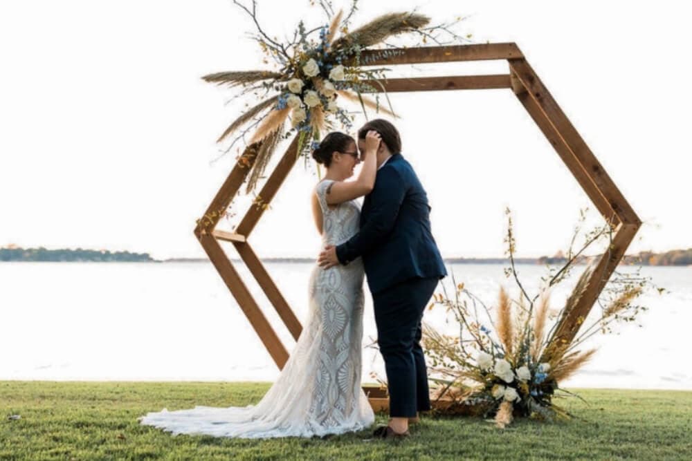A couple in wedding attire embraces under a floral arch with a lake in the background.