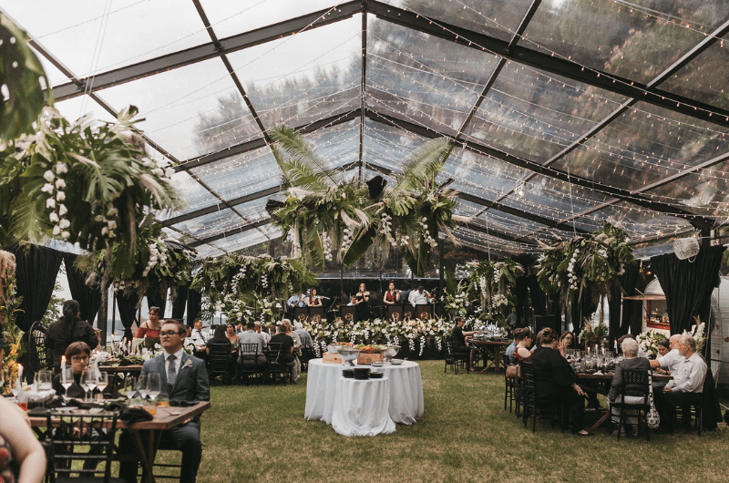 A beautifully decorated tented dining area filled with guests, lush greenery, and soft lighting.