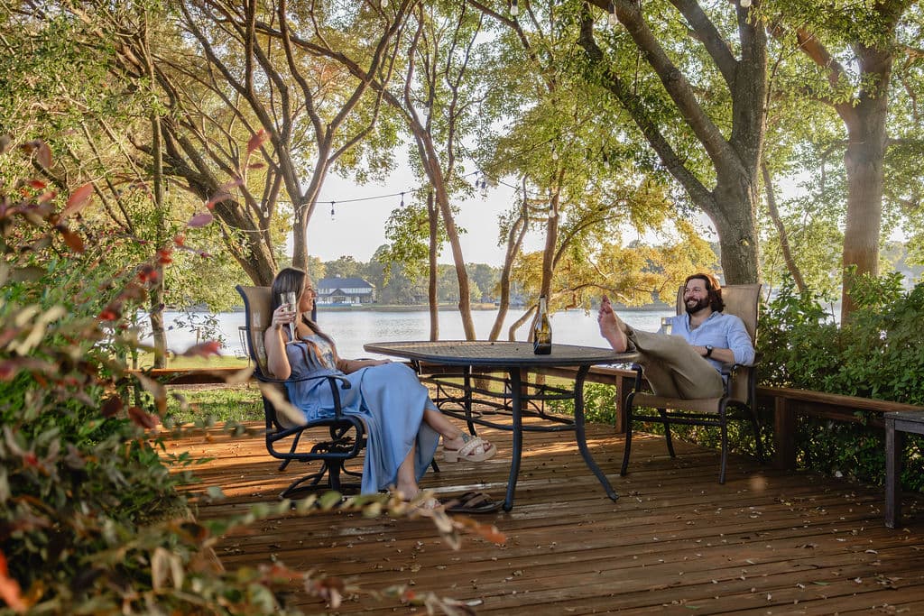 A woman in a blue dress and a man in a white shirt relax on a wooden deck by a lake, surrounded by trees. A woman in a blue dress and a man in a white shirt relax on a wooden deck by a lake, surrounded by trees.