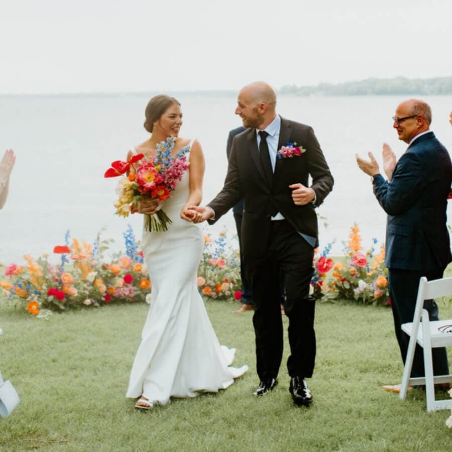 A bride and groom joyfully walk hand in hand after their wedding ceremony, surrounded by colorful flowers and applauding guests by a lakeside.