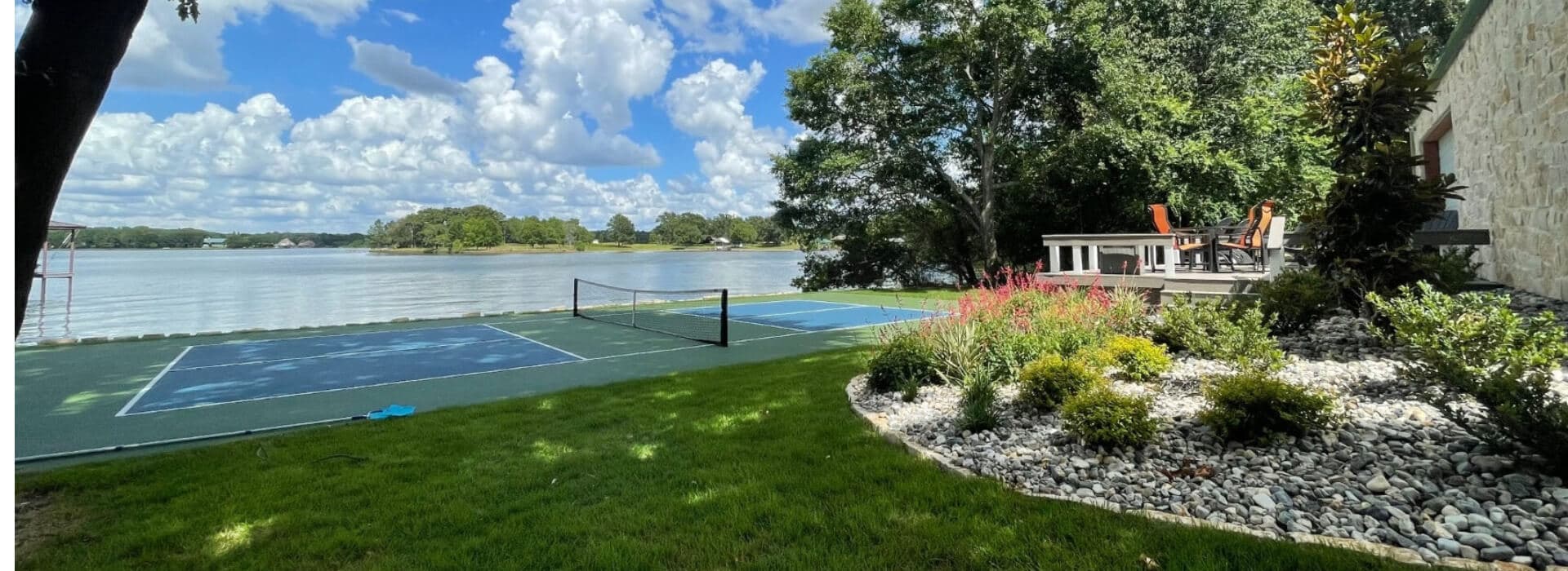 Lakeside view featuring a tennis court and a patio surrounded by greenery.