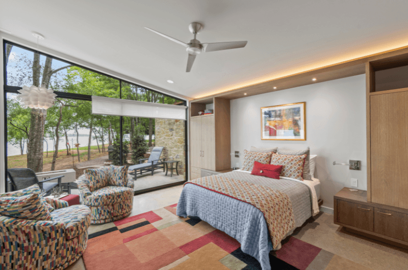 A modern bedroom features a bed with colorful bedding, two patterned chairs, and large windows overlooking a natural landscape.