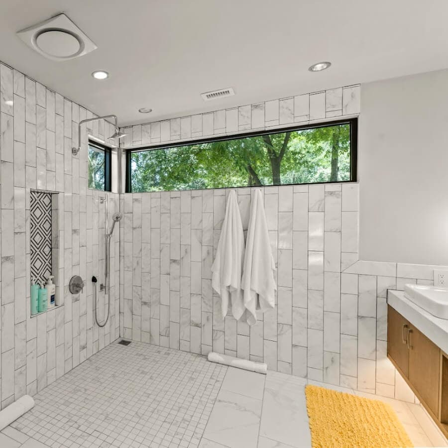 A modern bathroom featuring white tiled walls, a walk-in shower, and a window overlooking greenery.