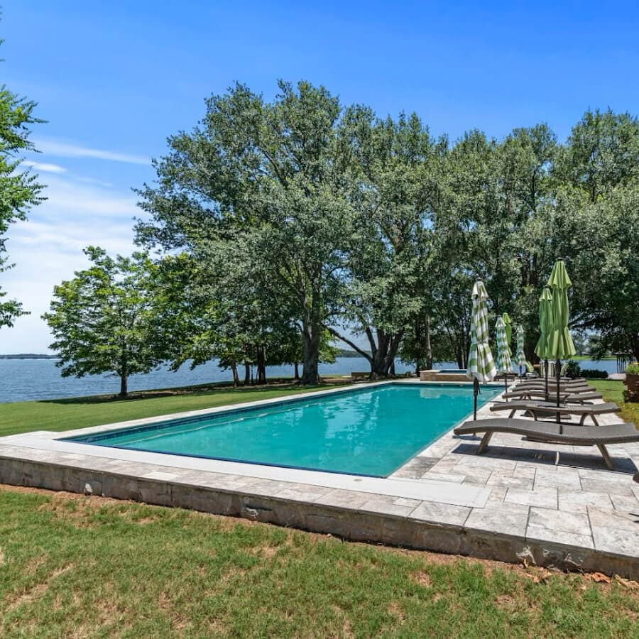 A serene swimming pool surrounded by trees and lounge chairs, with a lake in the background under a clear blue sky.