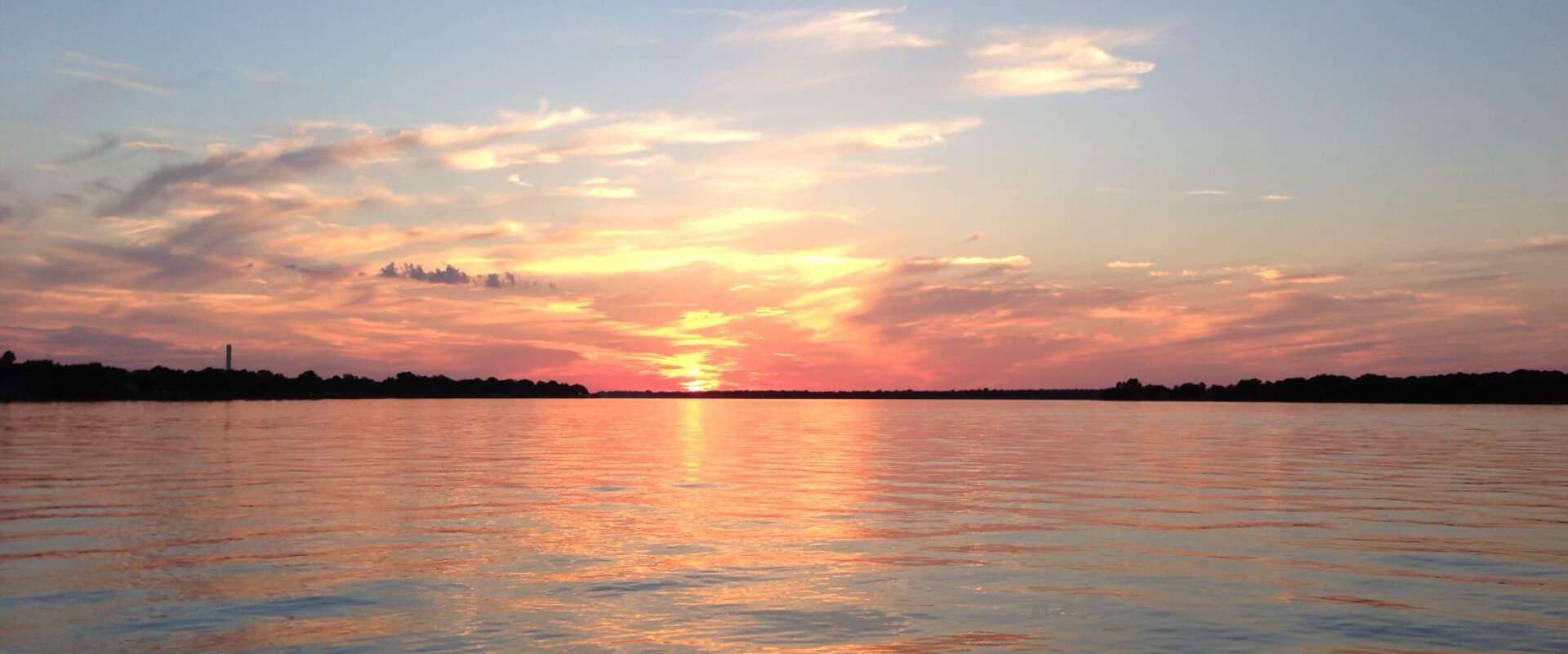 A vibrant sunset reflects on calm water, with clouds and a distant shoreline.