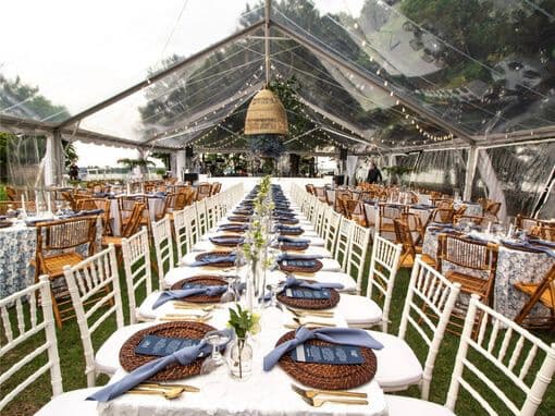 A beautifully arranged outdoor dining setup under a clear tent, featuring long tables with white chairs and elegant place settings.