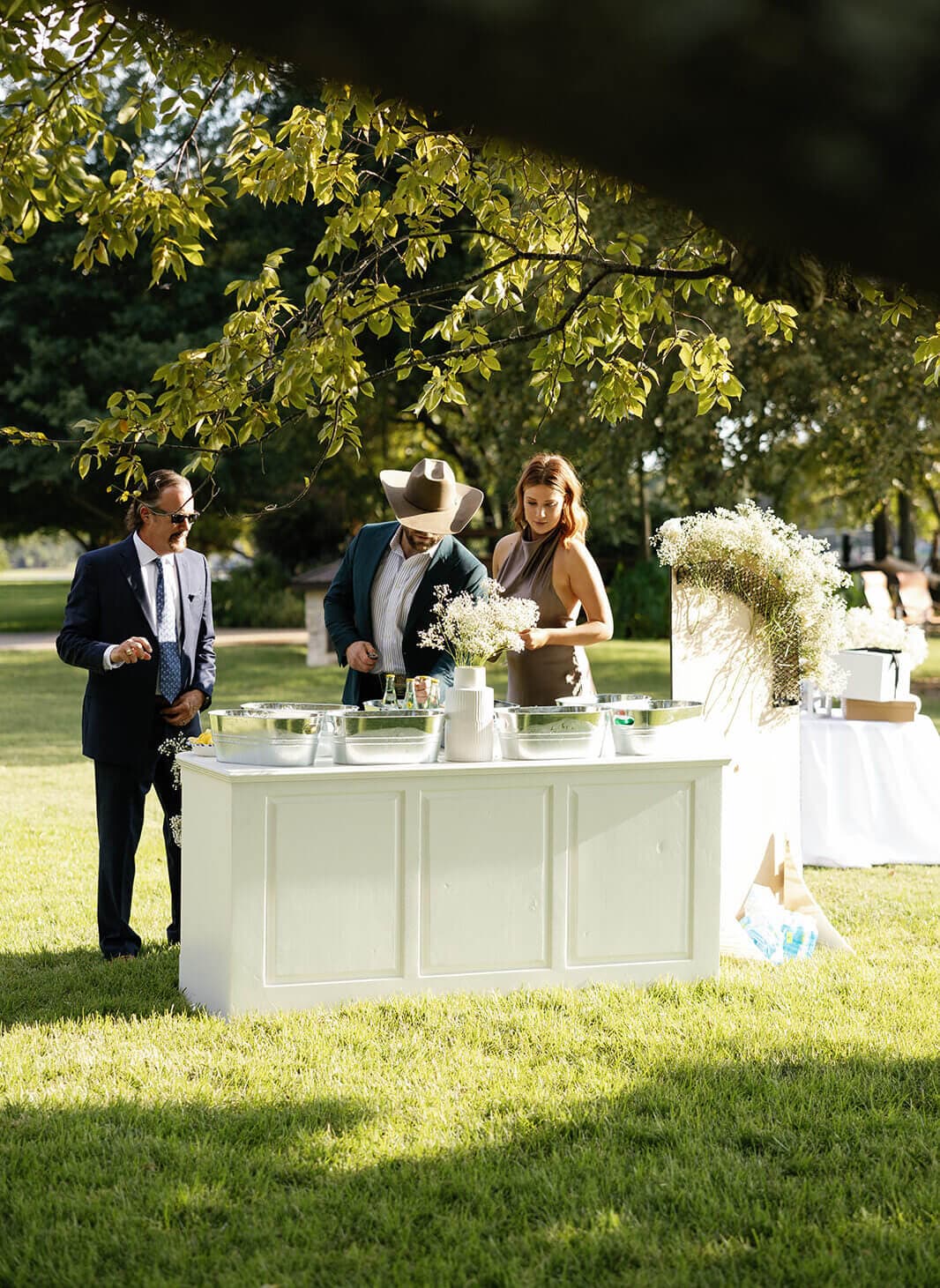 Three people gather around a white buffet table in a sunlit outdoor setting.