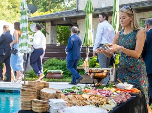 A woman prepares a plate from a buffet table filled with various foods while guests chat in the background.