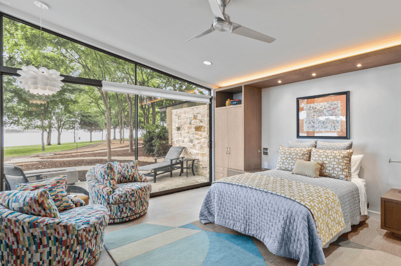 A modern bedroom with a large window view, featuring colorful chairs, a patterned bedspread, and natural light.