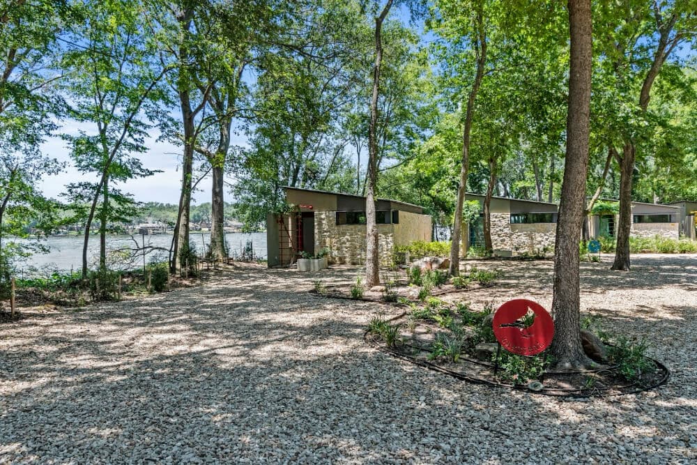 Two modern cabins nestled among trees by a riverbank, surrounded by gravel and greenery.
