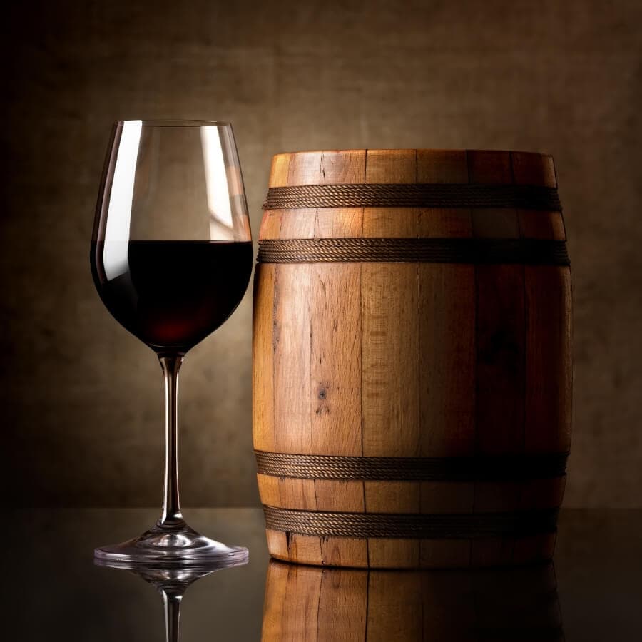 A glass of red wine beside a wooden barrel on a reflective surface.