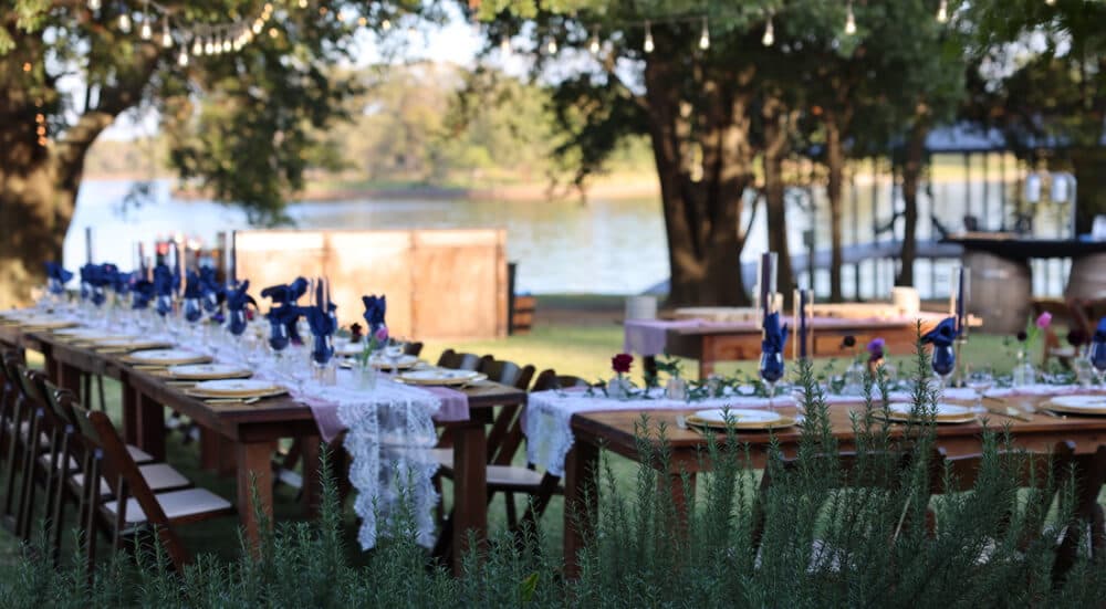 A beautifully arranged long outdoor dining table set near a lake, adorned with flowers and elegant tableware.